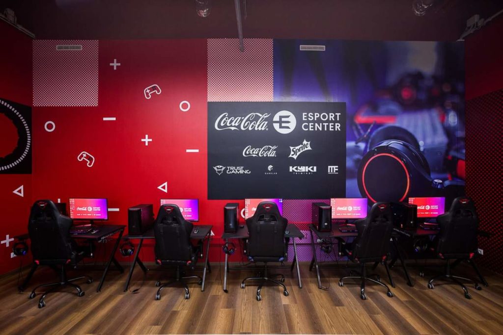 A képen a Play IT egyik szobája látható a falon szponzoráló cégek logóival. A szponzoráló cégek a következők: Coca-Cola, Esport center, Sprite, True Gaming, ITF, Kyiki